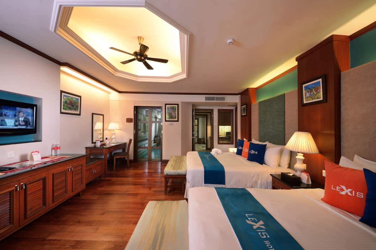 Habitación de hotel con dos camas, ventilador de techo, televisor, escritorio y baño visible a través de una puerta de cristal.