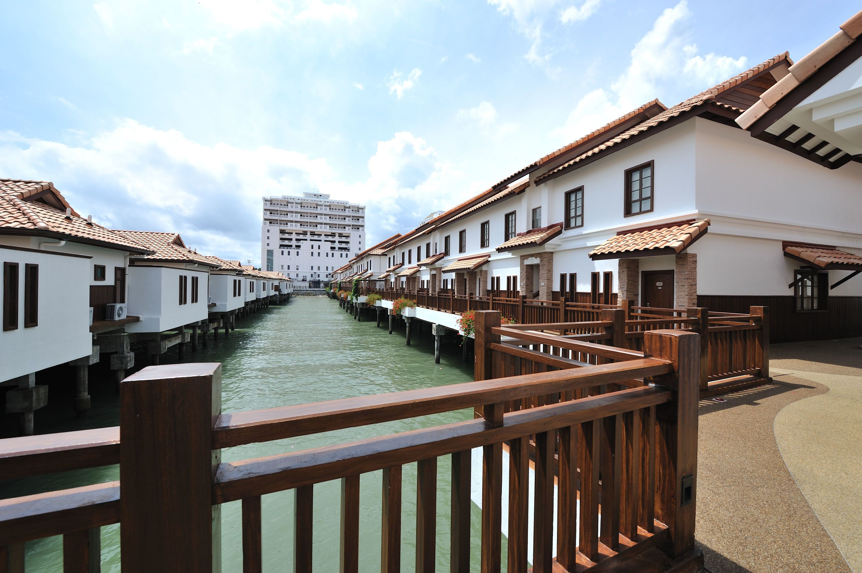 Un paseo junto al canal con barandas de madera, edificios blancos con tejados de azulejos y un cielo despejado.