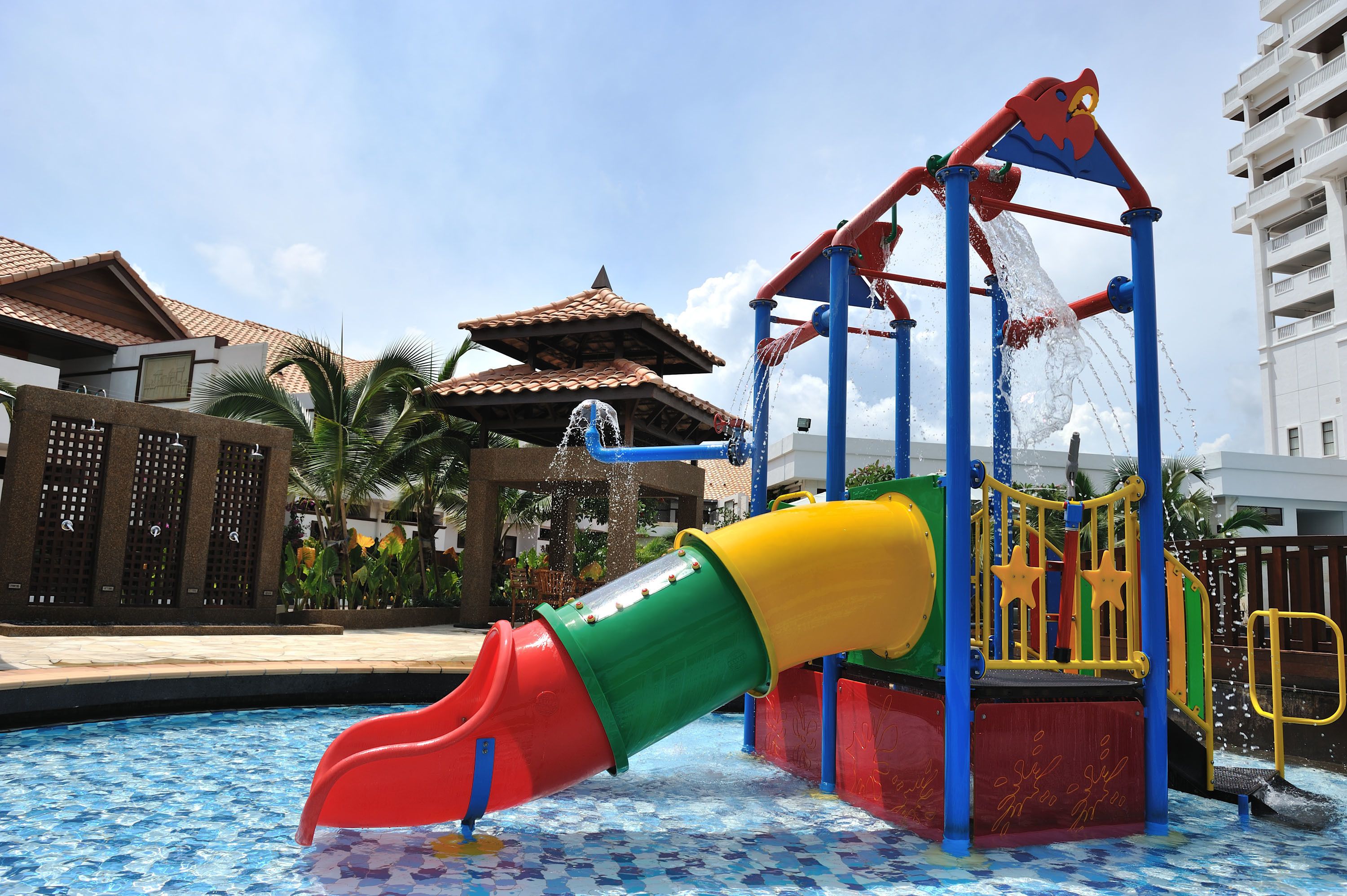 A water play area with a slide and climbing structure, surrounded by a pool and palm trees.