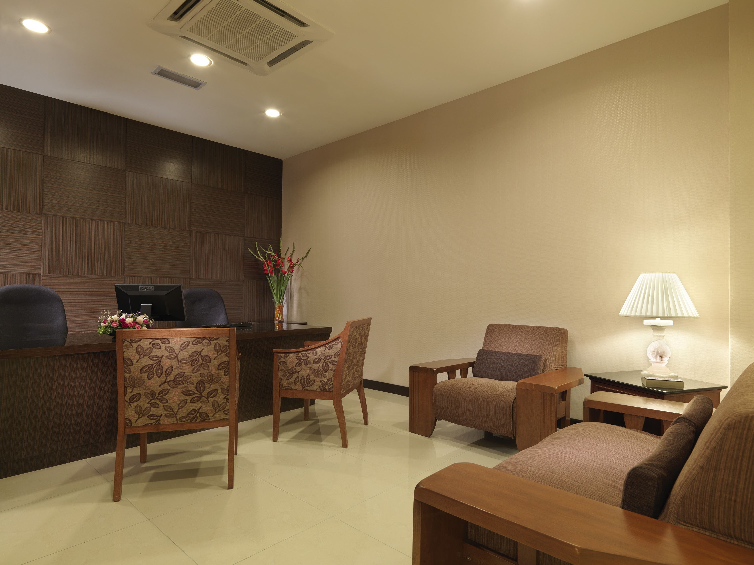 A hotel reception area with a wooden reception desk, two chairs, a sofa, a lamp, and a vase with flowers.