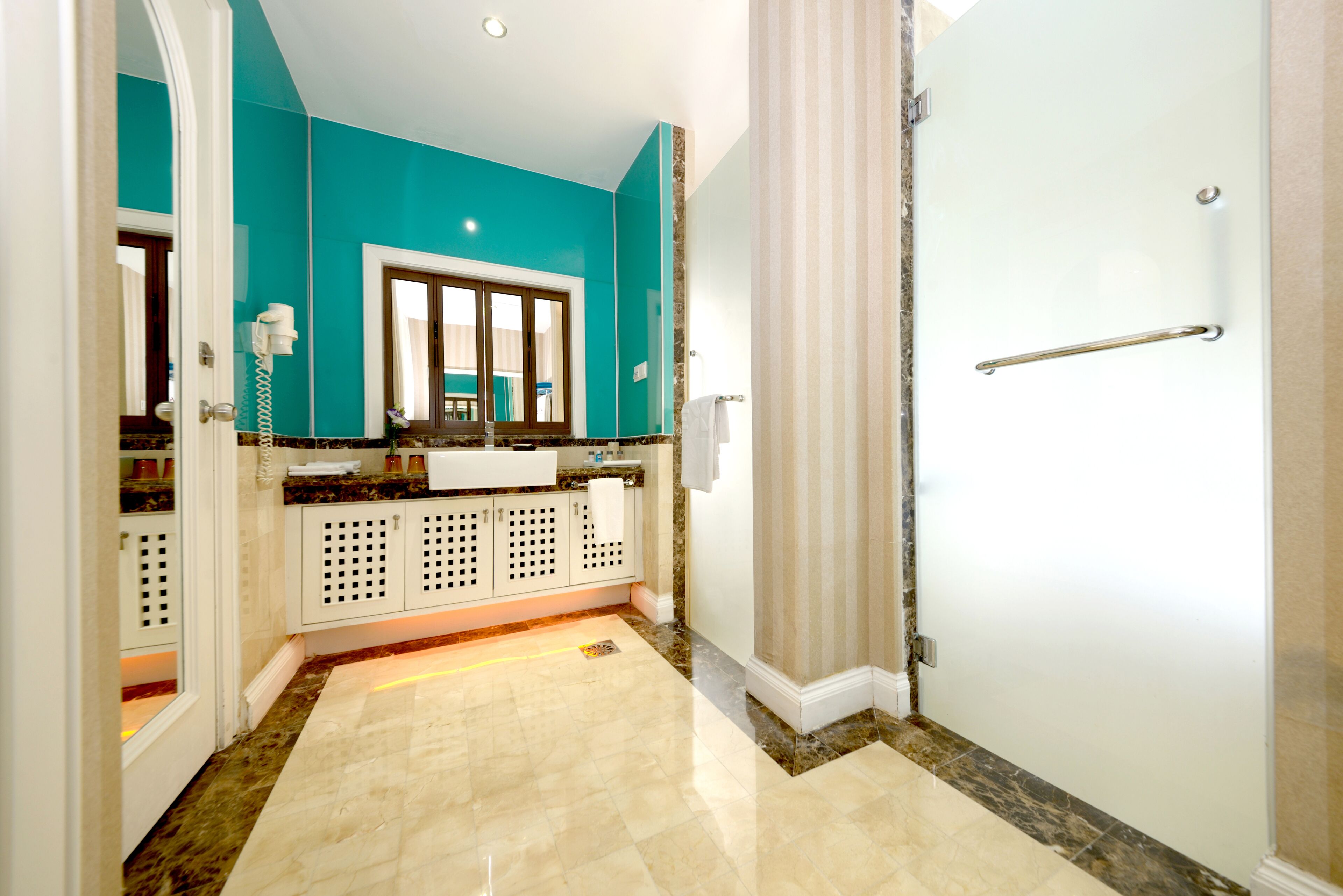 Un baño moderno con una pared de acento color turquesa, piso de mármol, un espejo grande y una ducha con puerta de cristal.