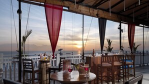 A terrace with wooden tables and chairs, a bar area, and a view of the sunset over the sea.