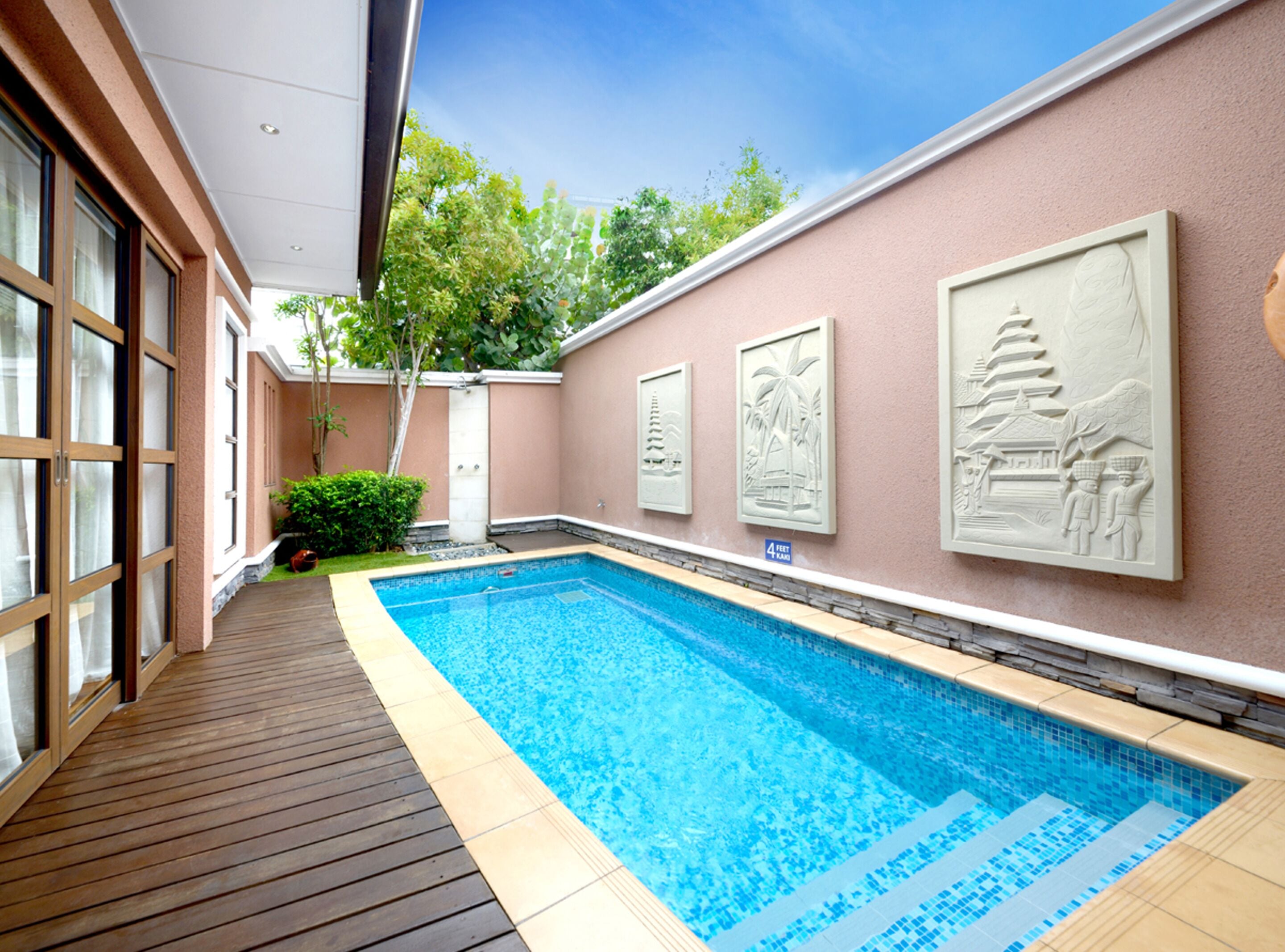 A swimming pool with a wooden deck, surrounded by a pink wall with decorative reliefs.