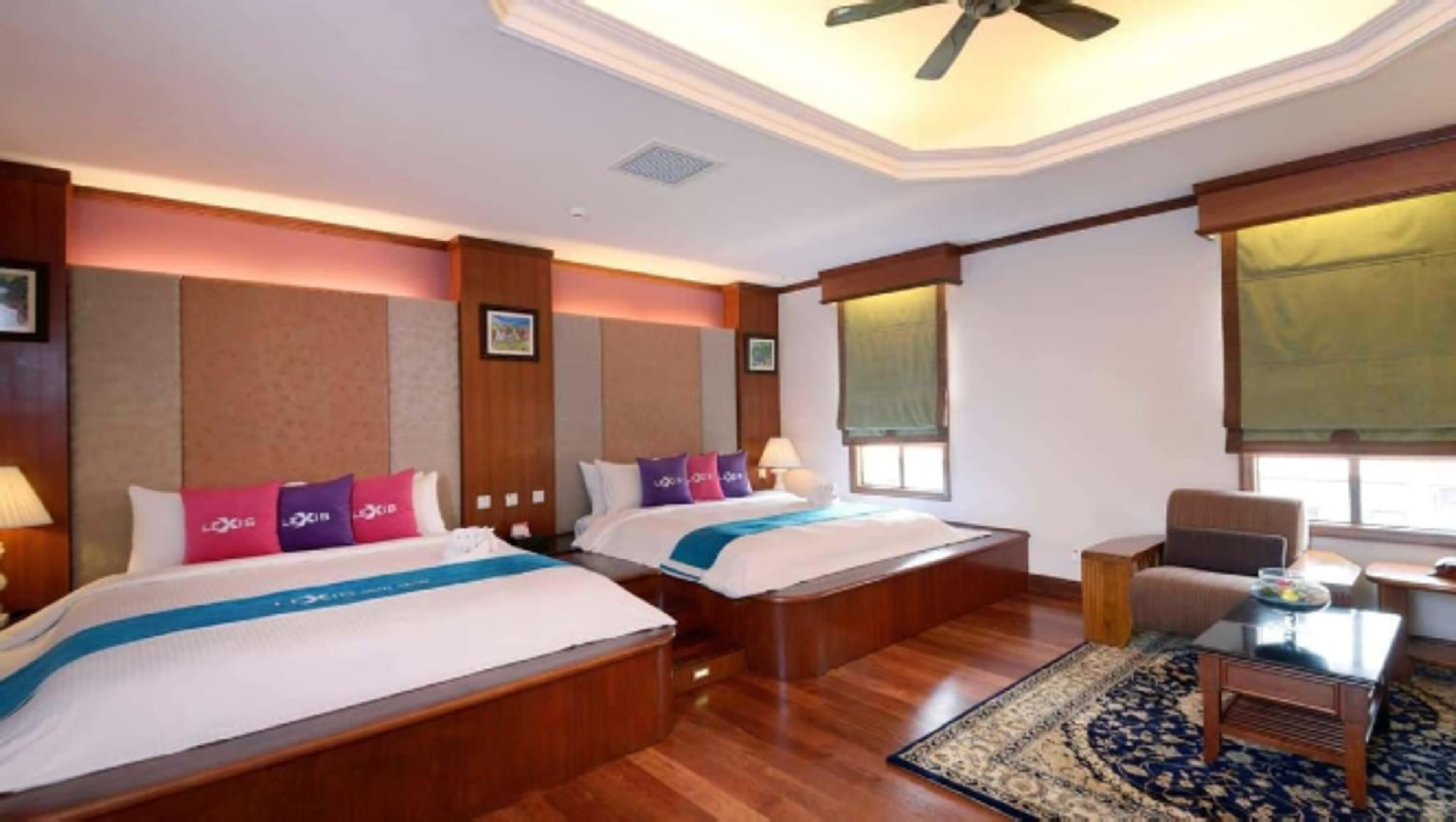 A hotel room with two beds, a desk, a chair, and a ceiling fan.