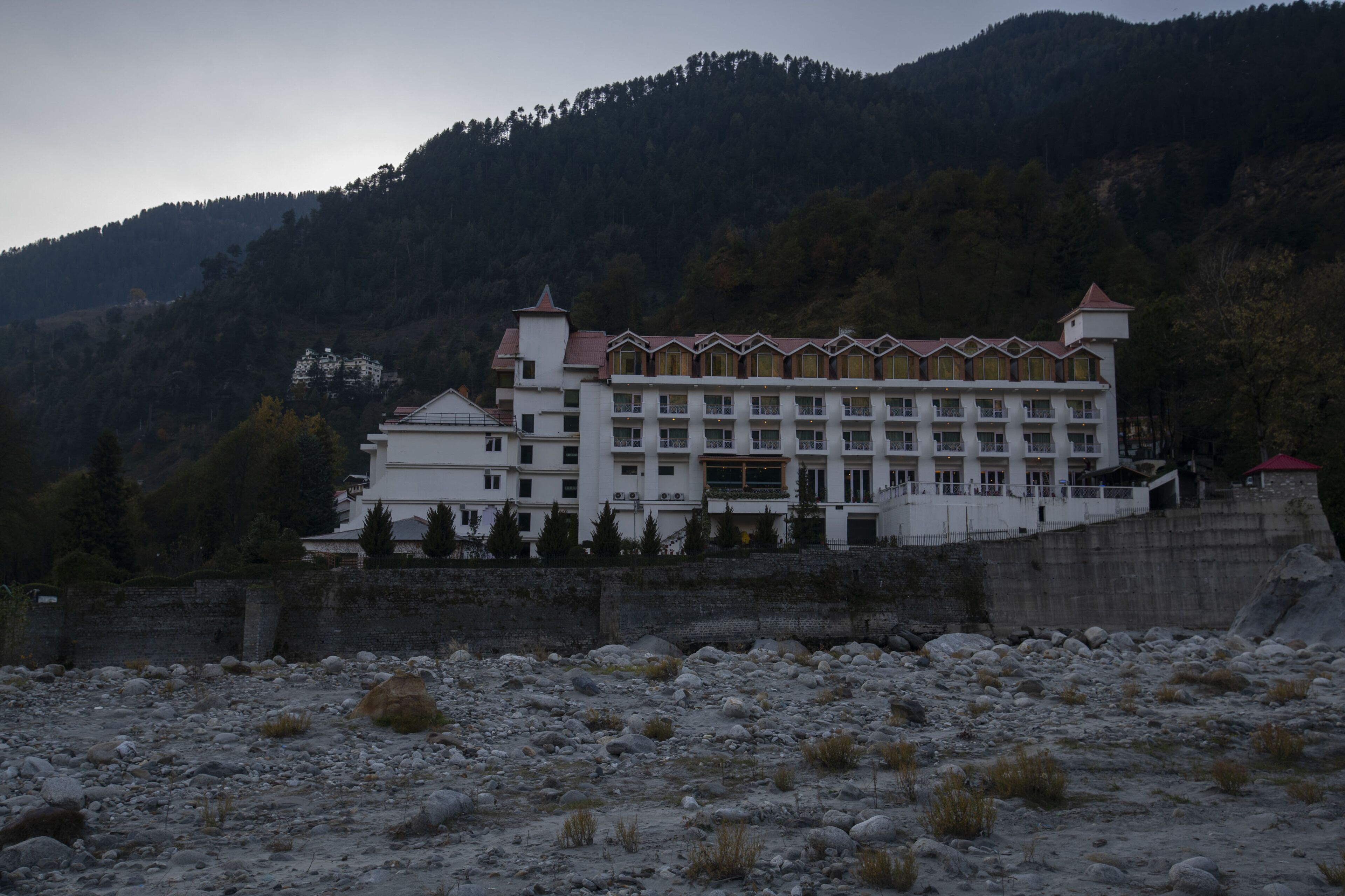 A large, multi-story building with a symmetrical facade, situated on a rocky terrain with a forested hill in the background.
