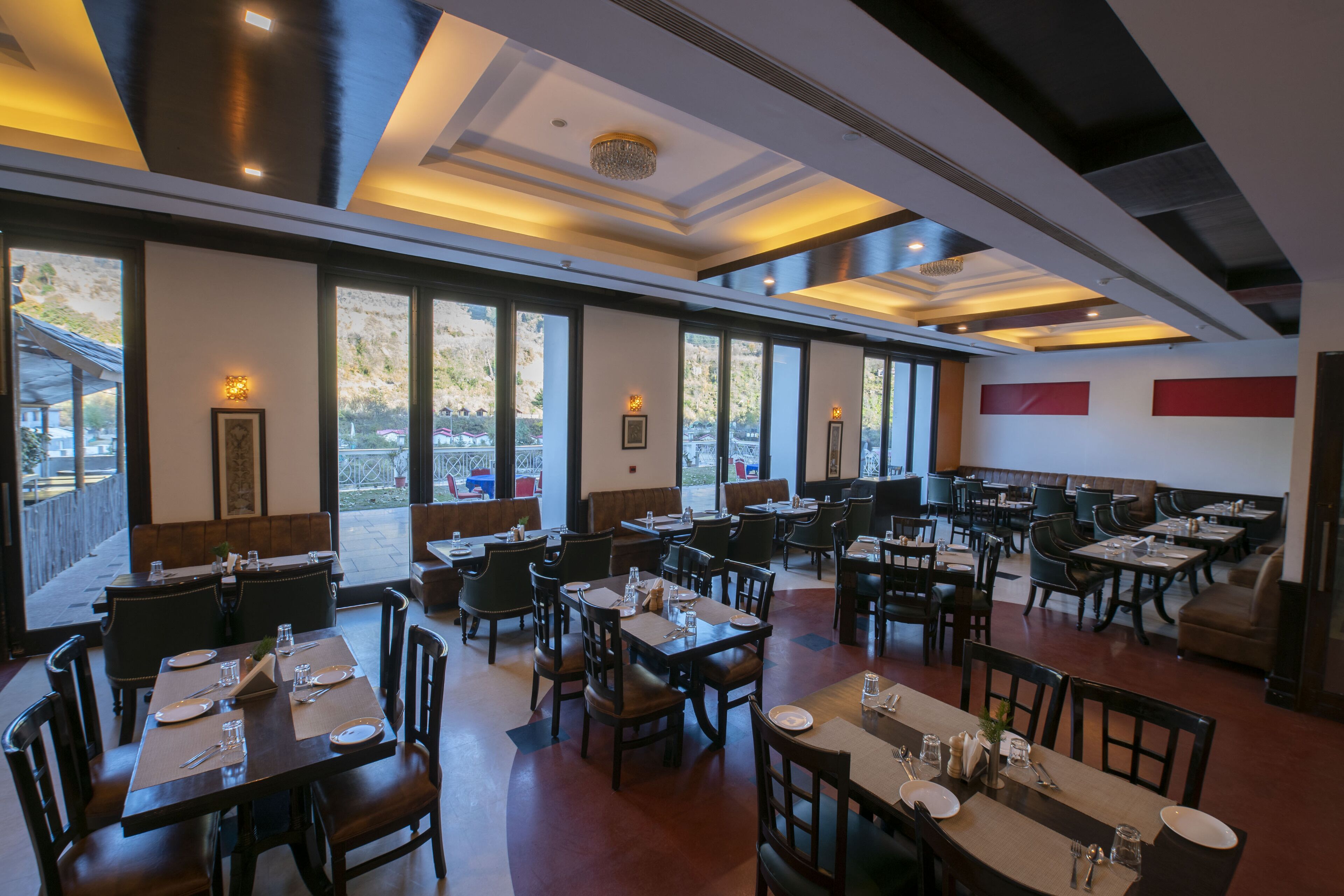 A spacious restaurant with multiple dining tables and chairs, large windows offering views of the outdoors, and a modern ceiling design.