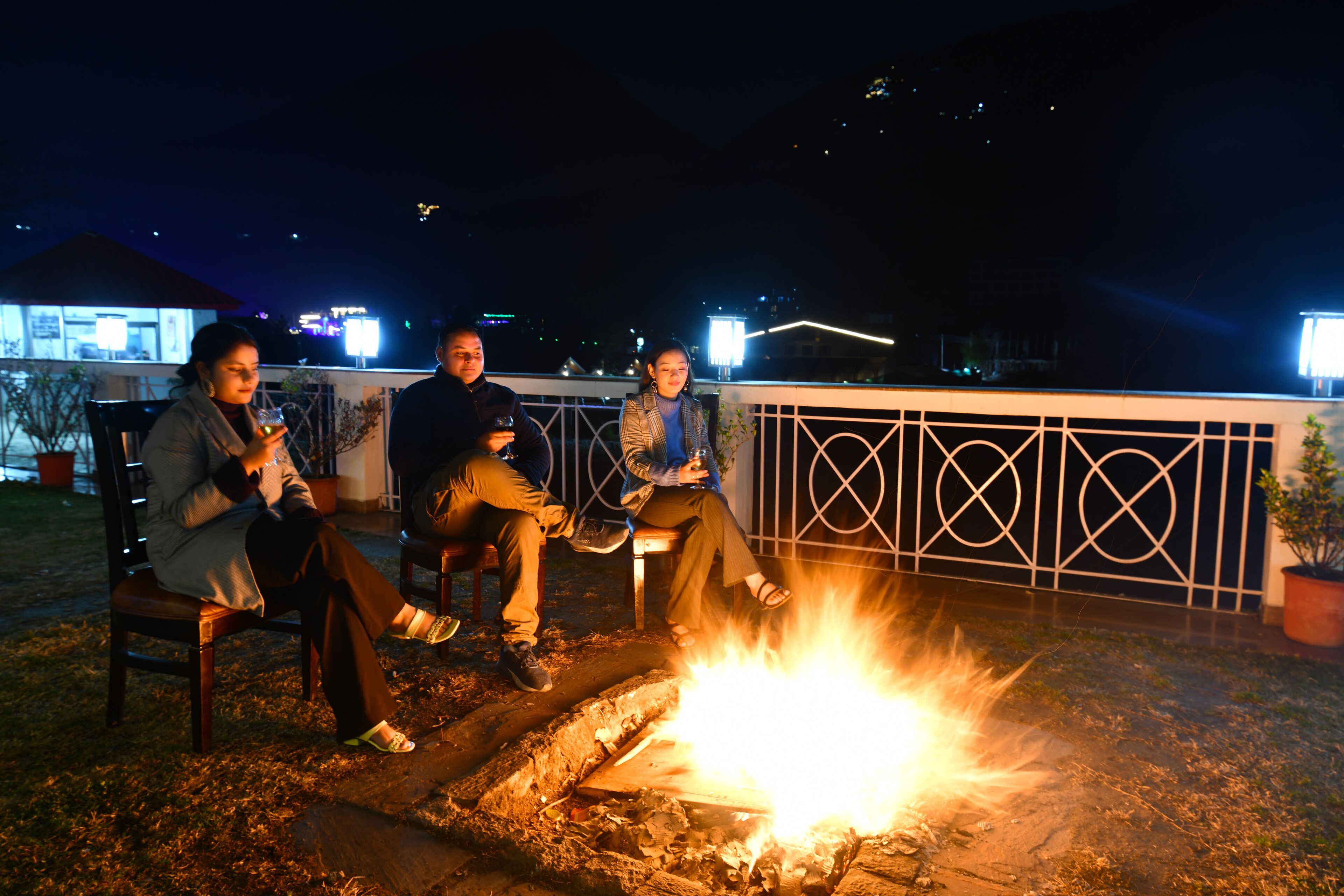 Tres personas sentadas junto a una fogata en una terraza en la azotea, con barandilla metálica y vista del paisaje urbano al fondo.