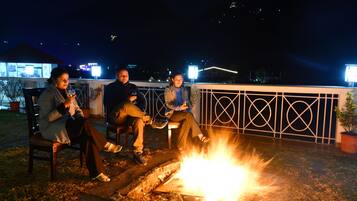 Tres personas sentadas junto a una fogata en una terraza en la azotea, con barandilla metálica y vista del paisaje urbano al fondo.