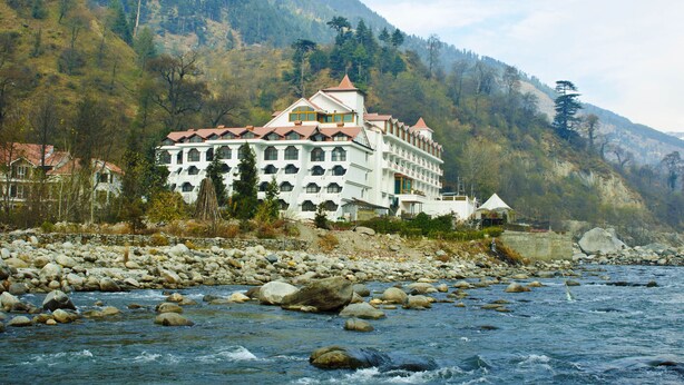 A large, multi-story hotel with a red roof, situated by a river with visible rocks and a mountainous backdrop. - Mastiff Grand Manali (Manali)