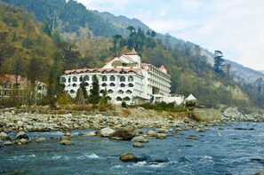 A large, multi-story hotel with a red roof, situated by a river with visible rocks and a mountainous backdrop. - Mastiff Grand Manali (Manali)