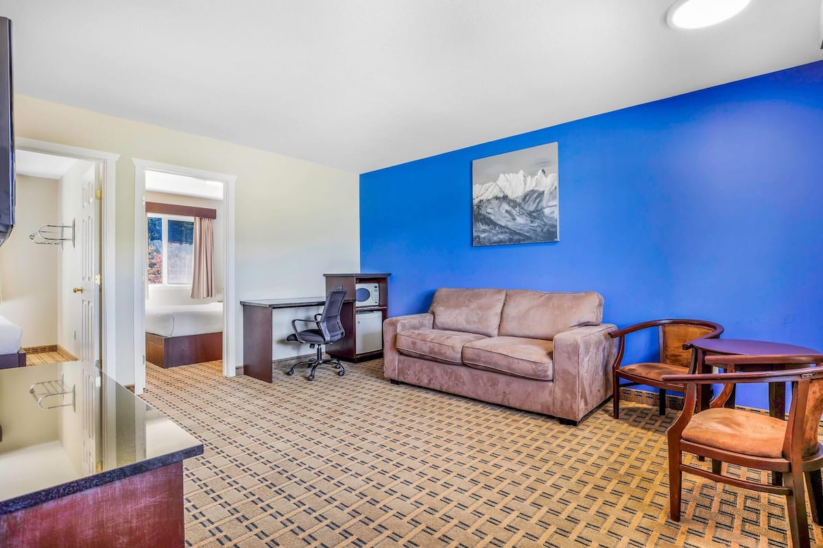 A hotel room with a patterned carpet, a beige sofa, a wooden coffee table, a chair, a desk with a computer, and a painting on the wall.