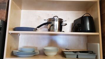 Un meuble de cuisine en bois, doté d’étagères où sont rangés une casserole en acier inoxydable, une bouilloire électrique noire, des assiettes blanches empilées, un saladier blanc et un plat de cuisson gris.