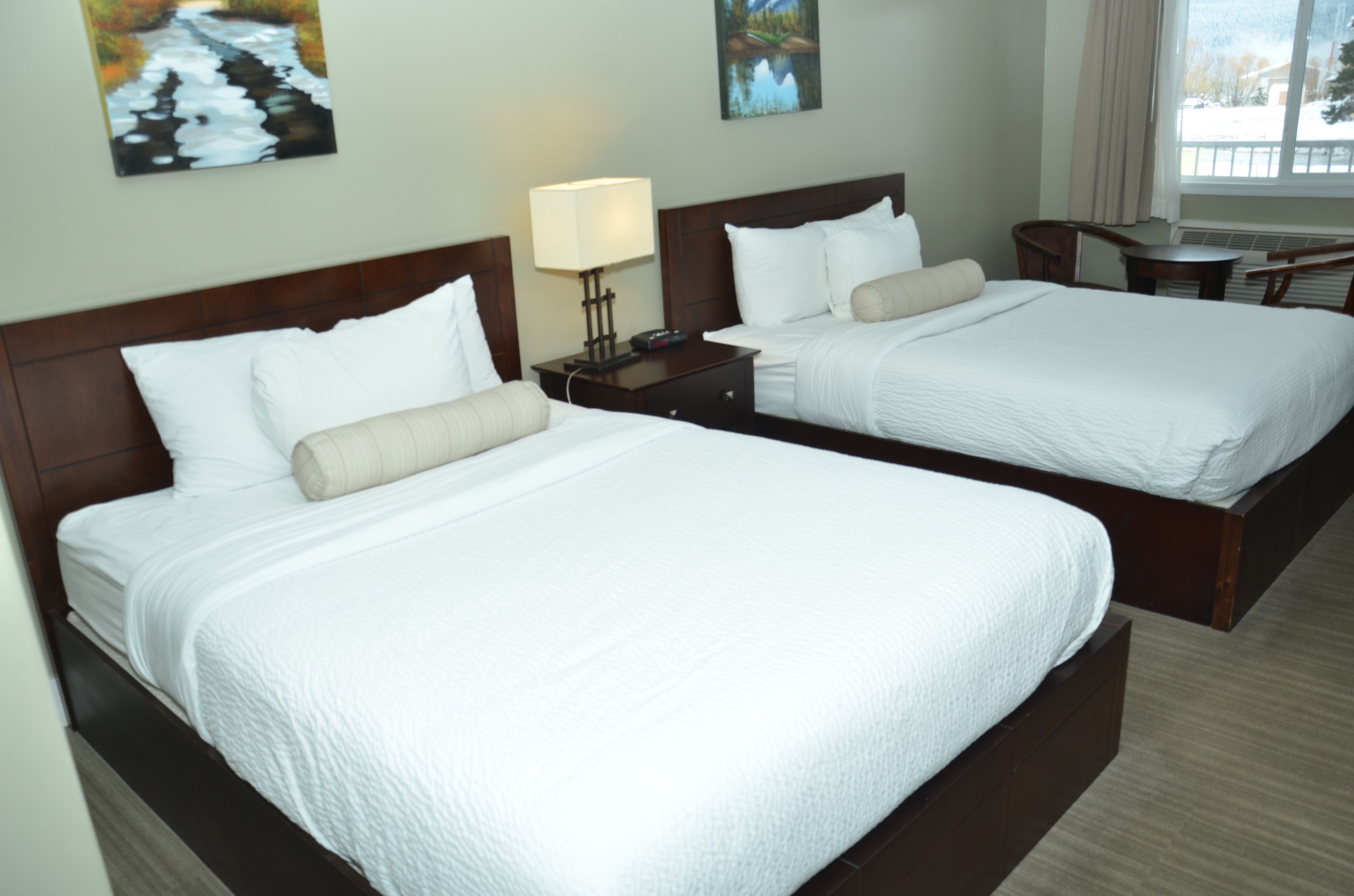 A hotel room with two beds, a nightstand with a lamp, and artwork on the walls.