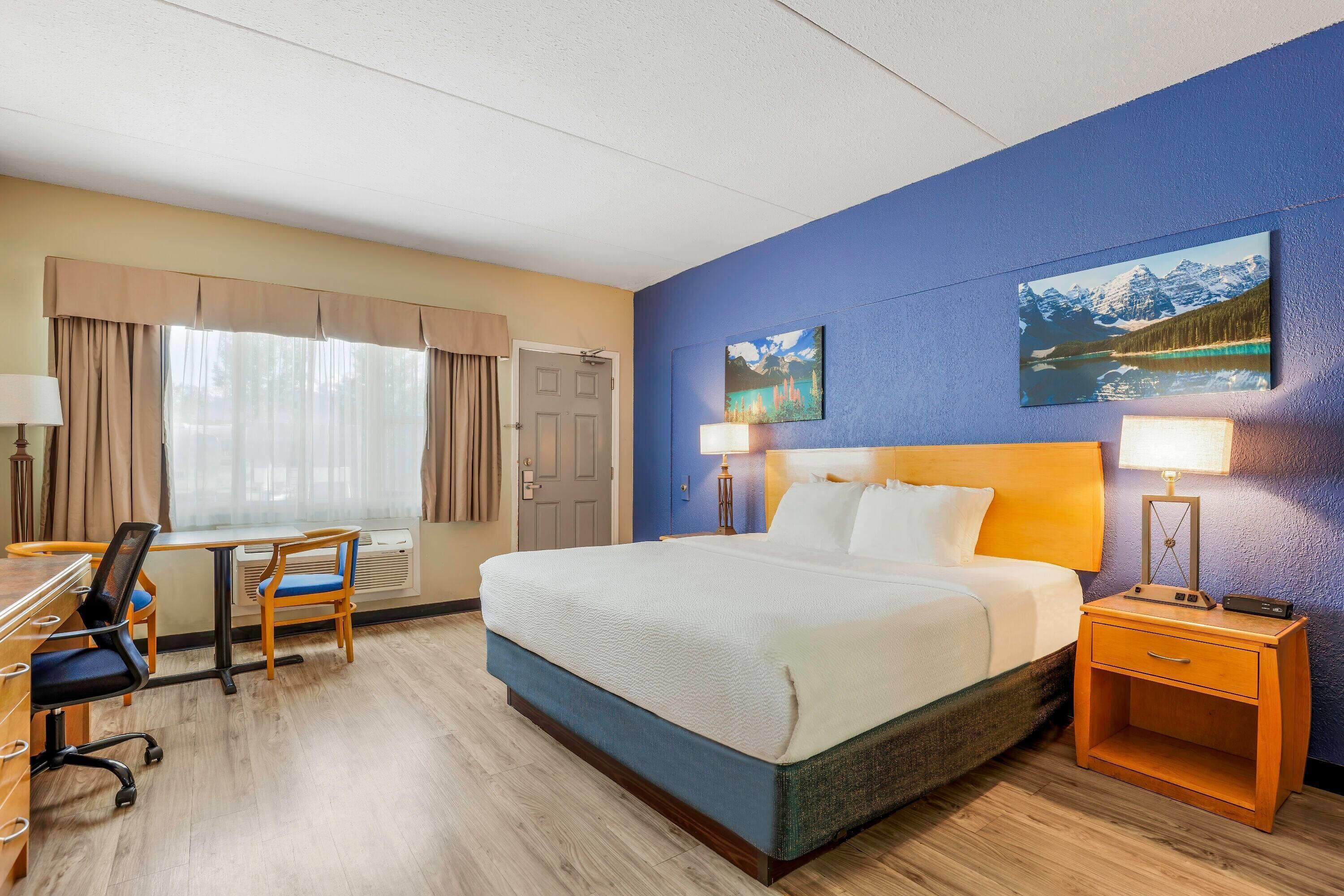 A hotel room with a bed, desk, chair, and two paintings on the wall.