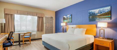 A hotel room with a bed, desk, chair, and two paintings on the wall.