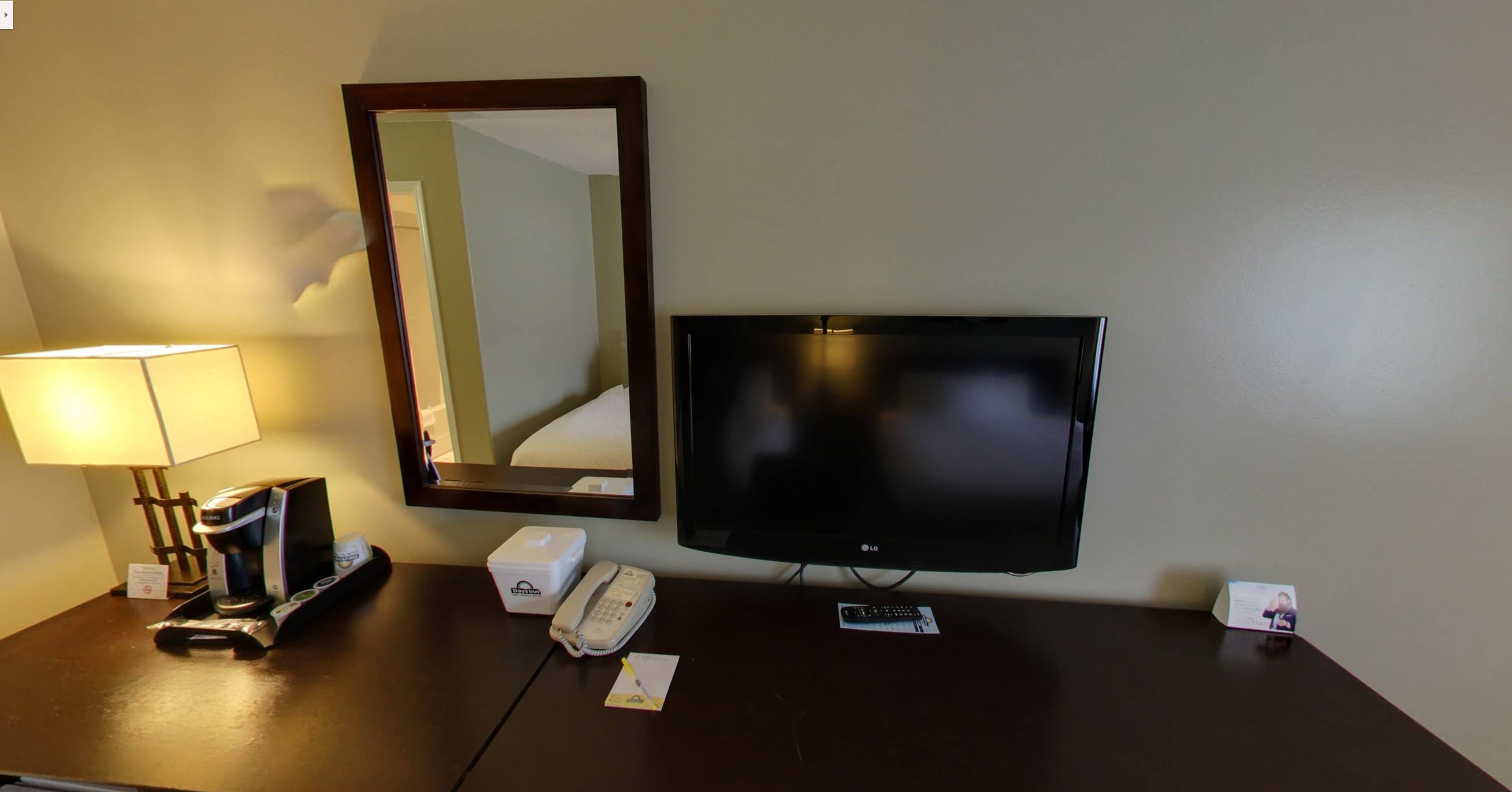 Habitación de hotel con televisor de pantalla plana, lámpara de noche, cafetera, teléfono y espejo sobre una cómoda de madera oscura.