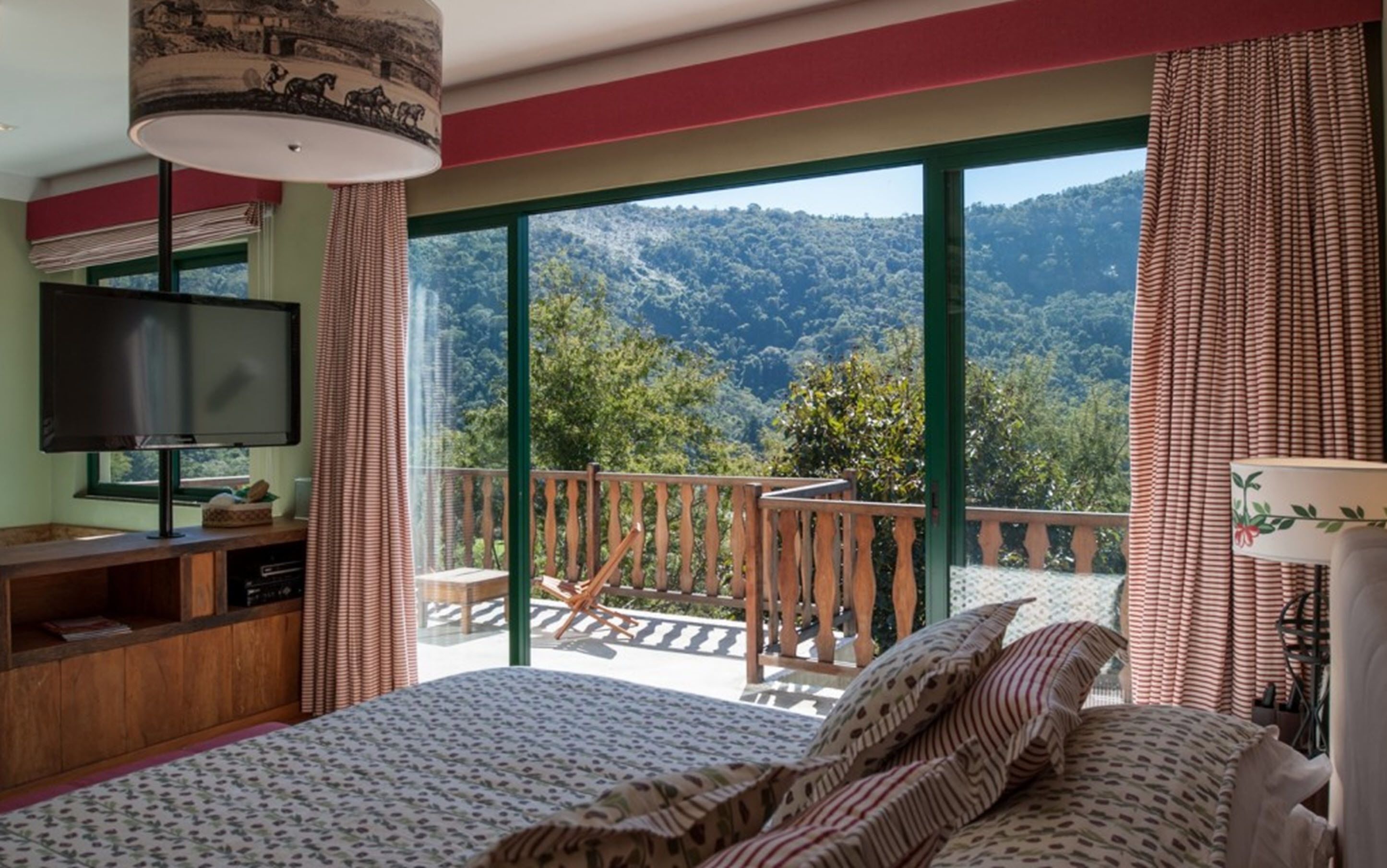 A bedroom with a large bed, a TV, and a balcony with a view of a forested mountain.