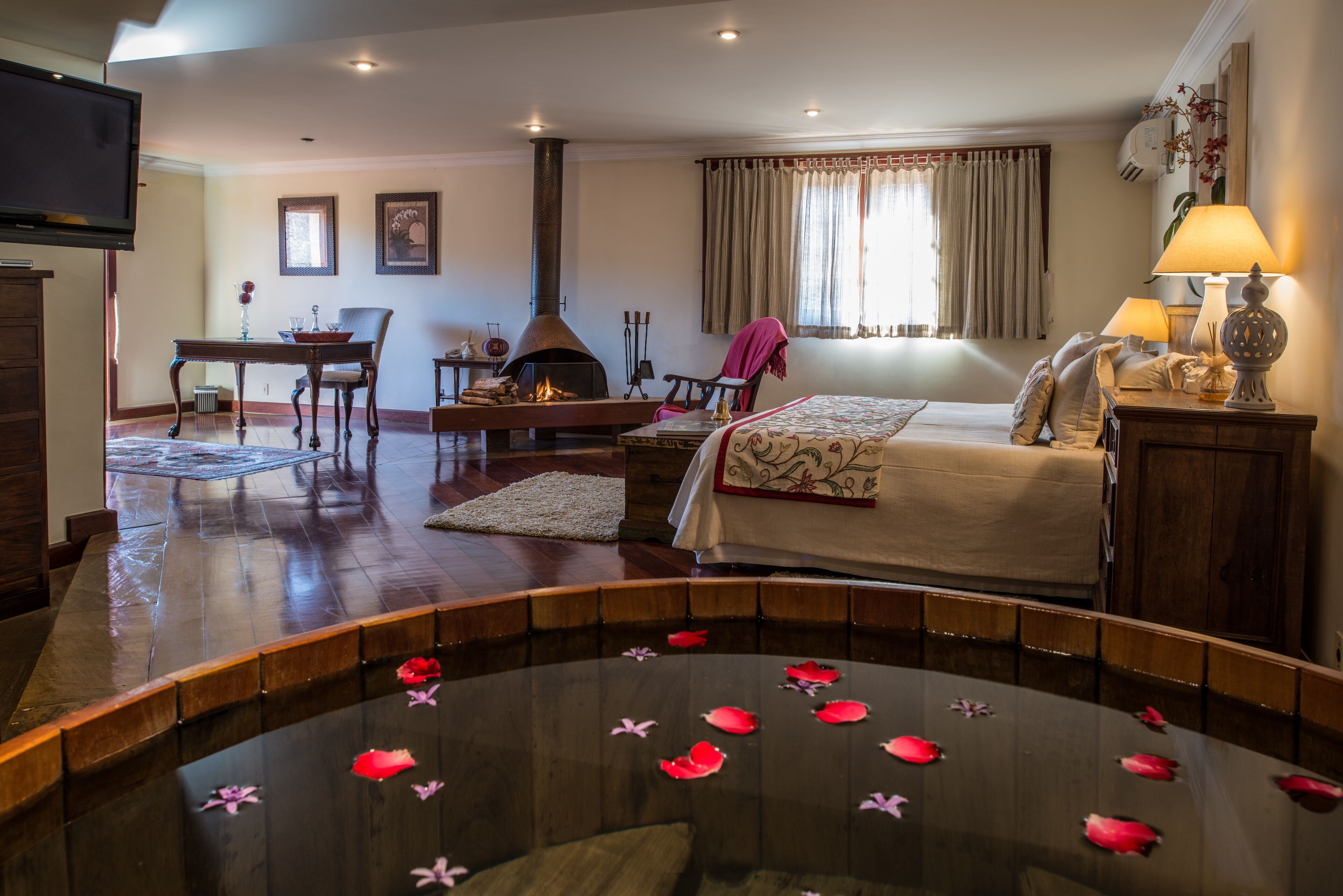 Une chambre d’hôtel avec un lit, une petite table sur laquelle est posé un vase, une cheminée et une piscine agrémentée de fleurs flottantes.