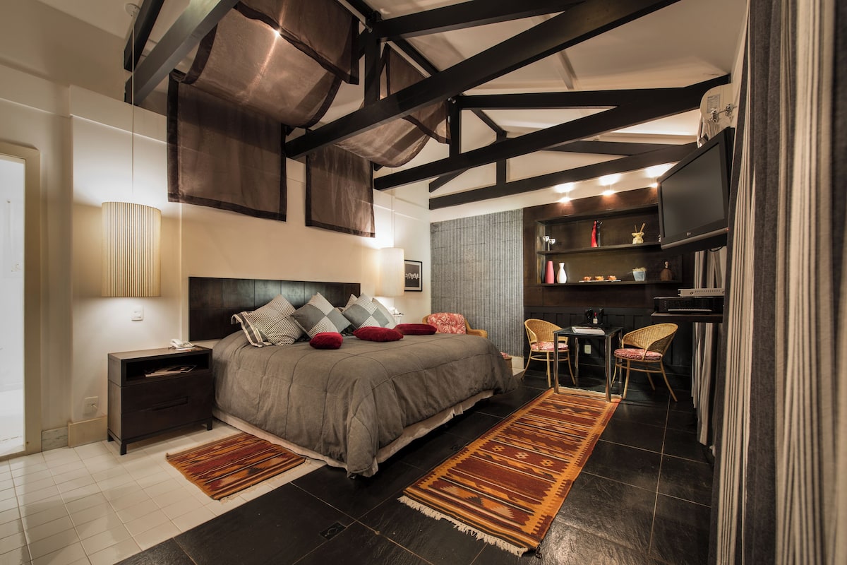 Ein modernes Schlafzimmer mit einem großen Bett, dunklen Holzbalken, einem Fernseher und einer kleinen Sitzecke.