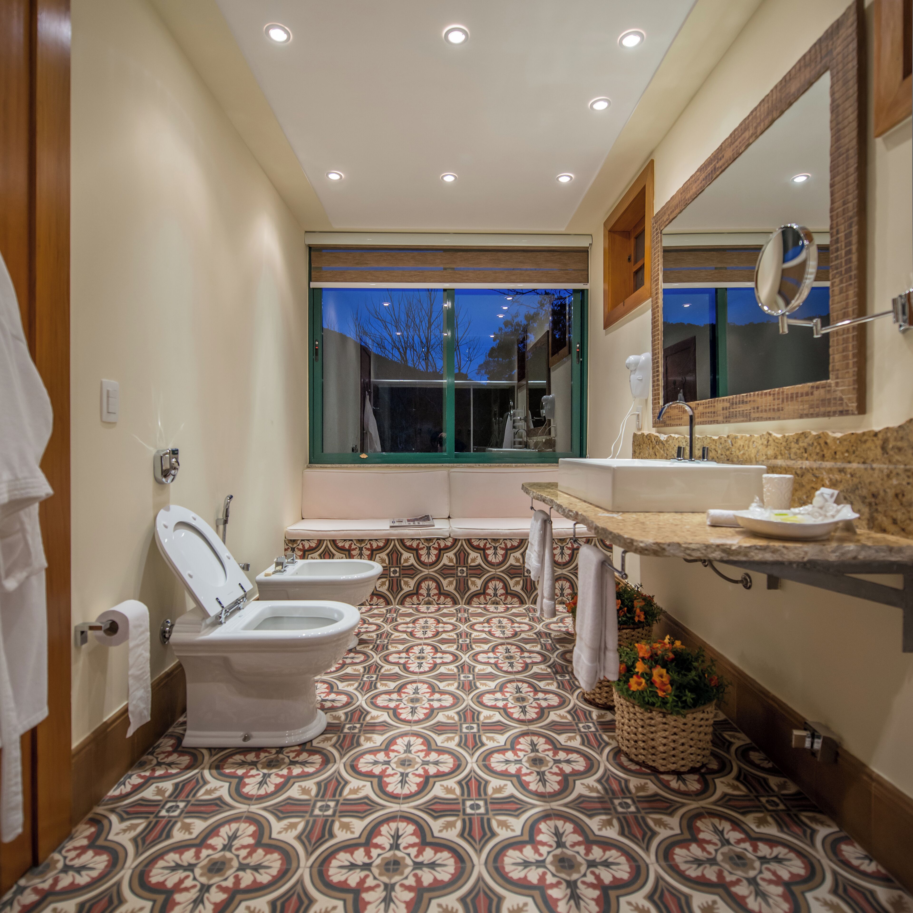 A bathroom with a patterned floor, a toilet, bidets, a sink, and a mirror.
