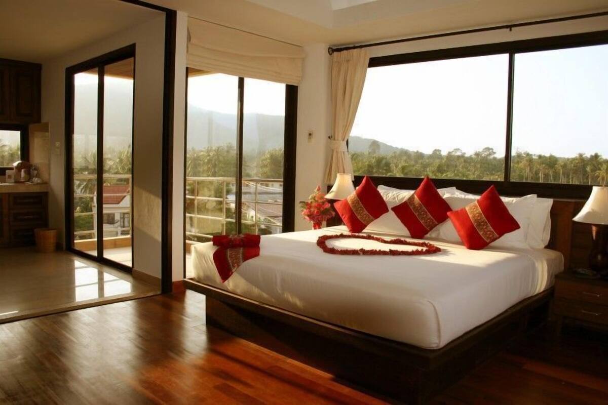 A large bed with red pillows and a heart-shaped decoration, a nightstand with a lamp, and a view of greenery through large windows.