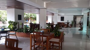 Amplo espaço interno com mesas e cadeiras de madeira, cercado por plantas em vasos.