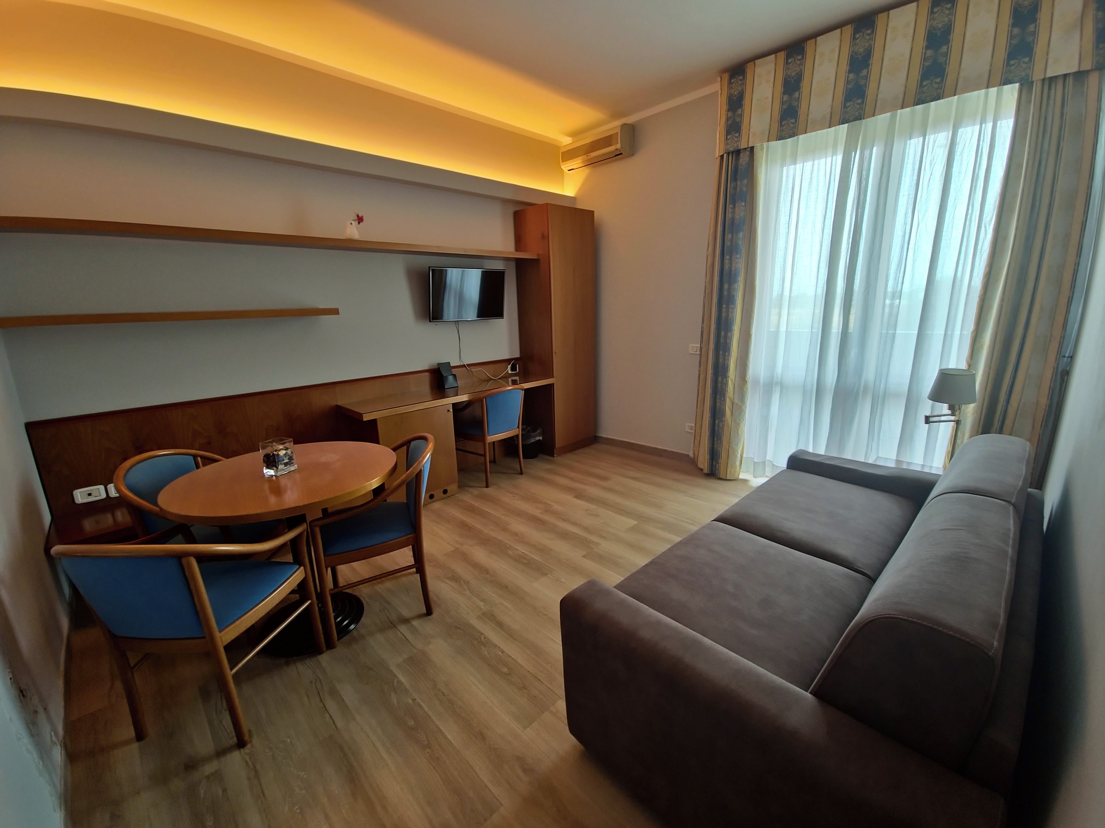 Een hotelkamer met een bank, een eettafel met stoelen, een aan de muur bevestigde televisie en een raam met gordijnen.