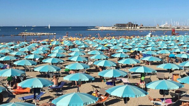 A beach with numerous light blue umbrellas and sun loungers, a clear sky, and a distant view of boats and buildings.