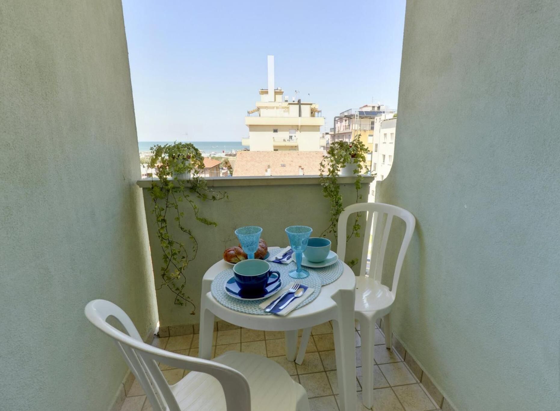 Un pequeño balcón con una mesa para dos, sillas blancas y vistas a edificios y al mar.