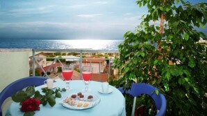 A table with a blue tablecloth, two glasses of red drink, a white cup, and a plate of pastries on a balcony overlooking the sea.