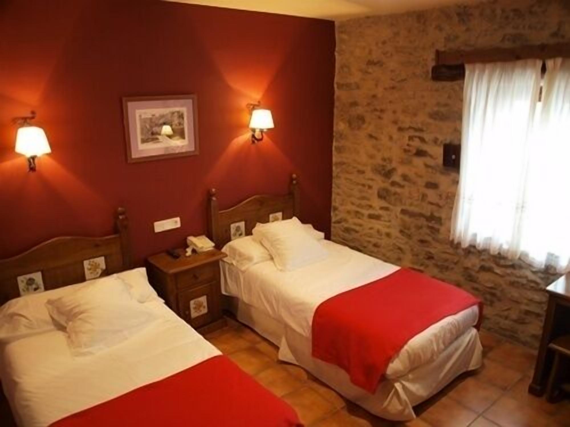 A room with two beds, each with a red blanket, a wooden headboard, and a nightstand with a lamp. There is a stone wall and a window with curtains.