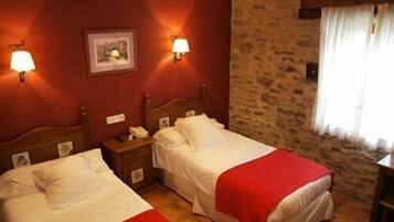 A room with two beds, each with a red blanket, a wooden headboard, and a nightstand with a lamp. There is a stone wall and a window with curtains.