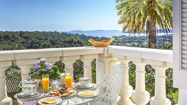 A balcony with a table set for breakfast, overlooking a scenic view of the sea and palm trees.