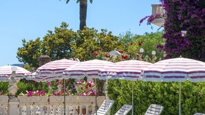 A poolside area with white lounge chairs and striped umbrellas, surrounded by lush greenery and flowering plants.