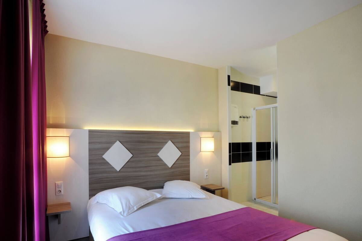 A hotel room with a bed, two bedside lamps, a nightstand, a mirror, and a shower area.
