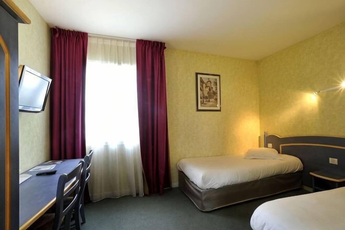 A hotel room with a bed, a desk with a chair, a television, and a window with curtains.