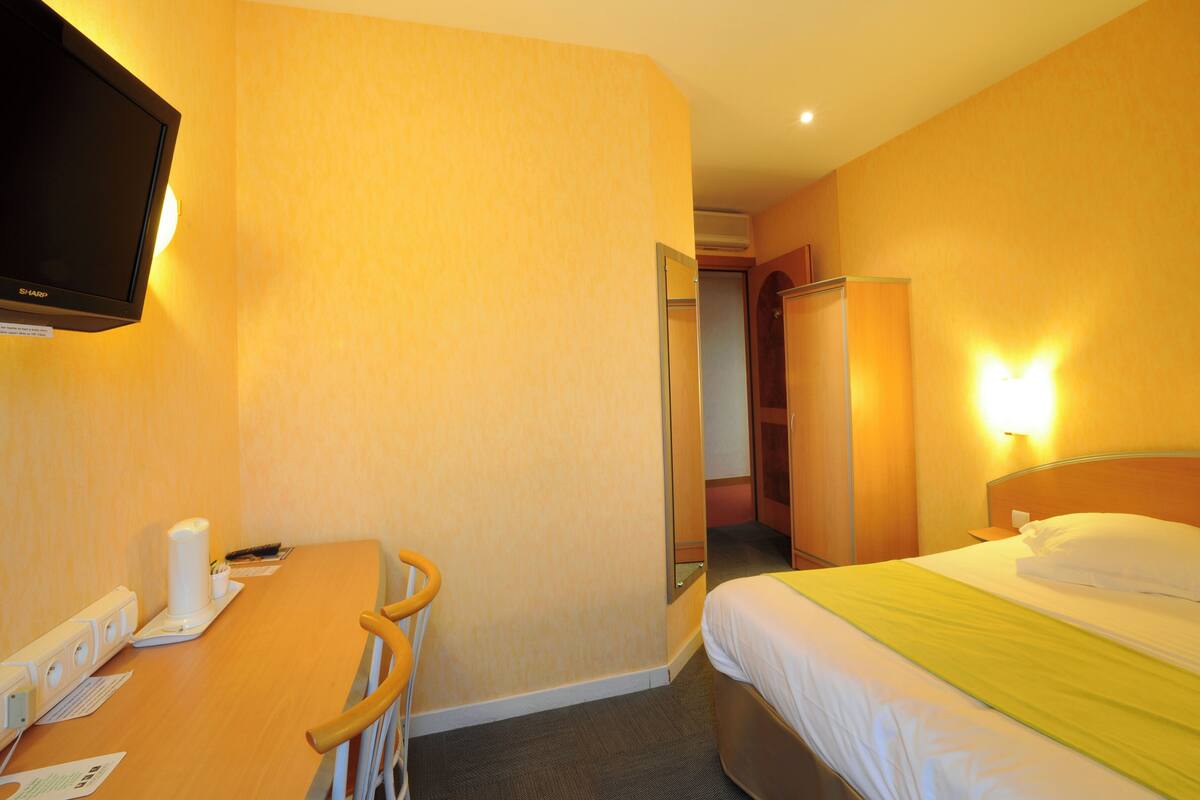 A hotel room with a bed, a desk with a chair, a television, and a bathroom visible through an open door.