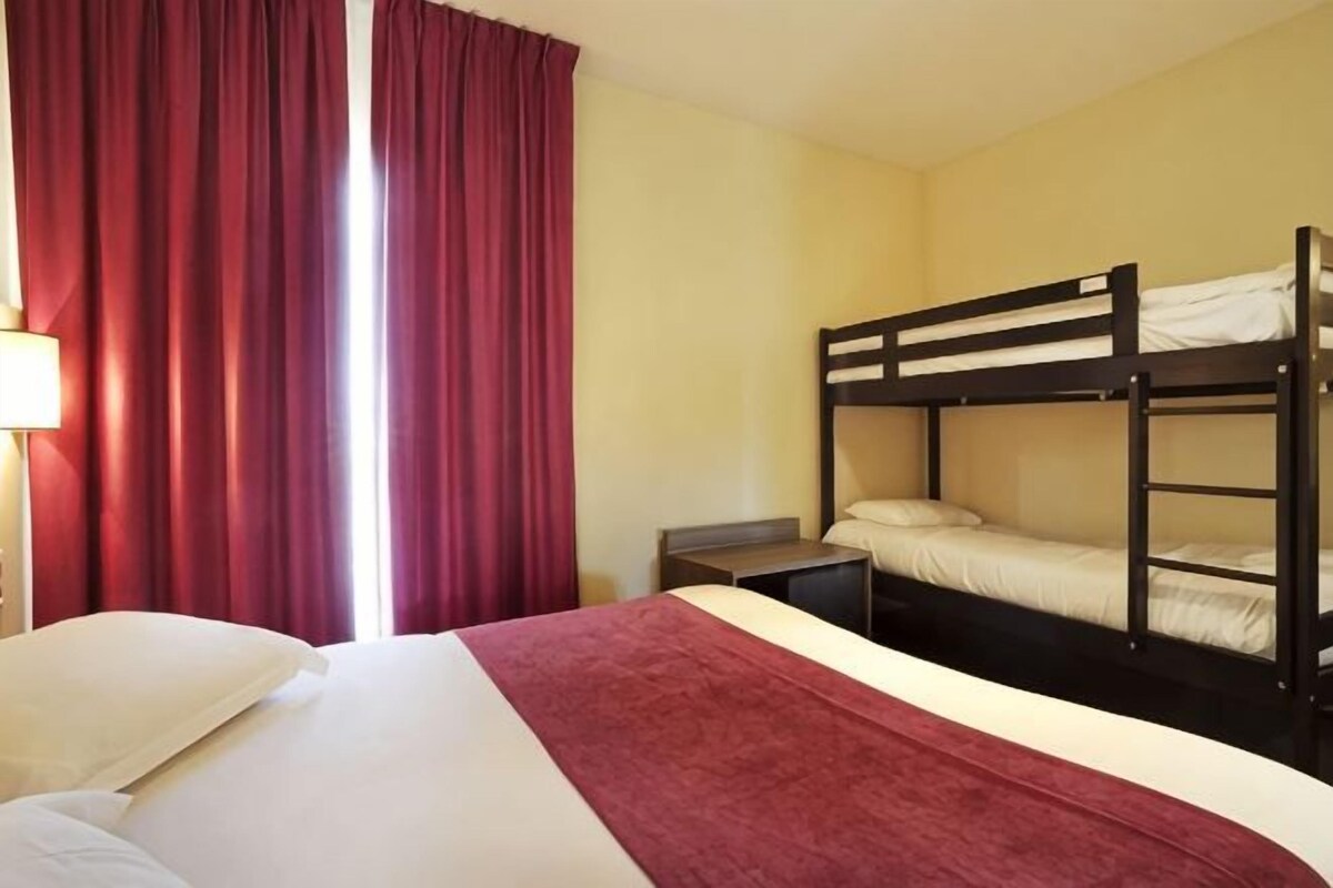 Ein Hotelzimmer mit Etagenbett, einem Einzelbett, einem Nachttisch und einem Fenster mit roten Vorhängen.