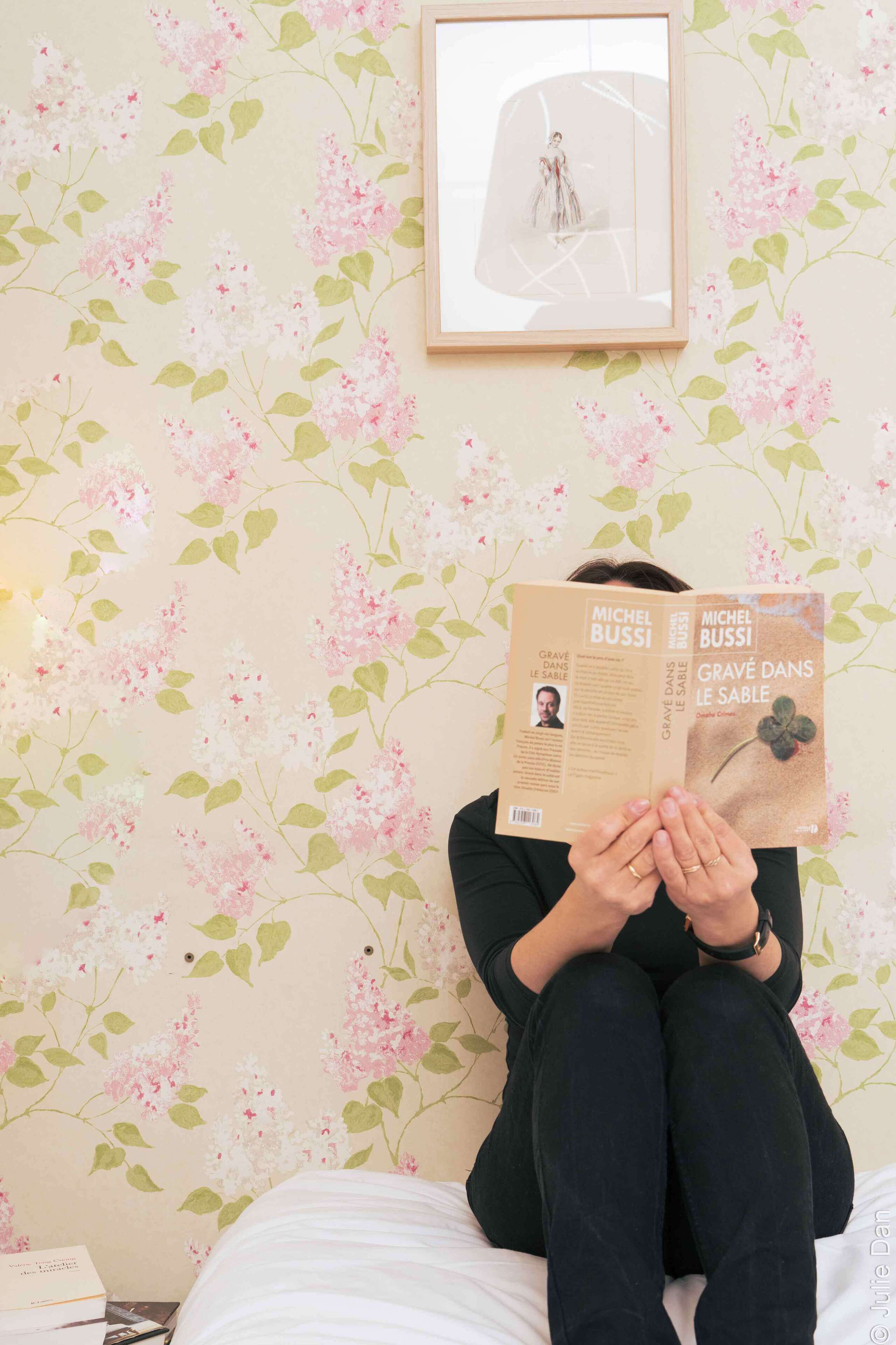 A person sitting on a bed with a book titled 'Grave dans le sable' by Michel Bussi.