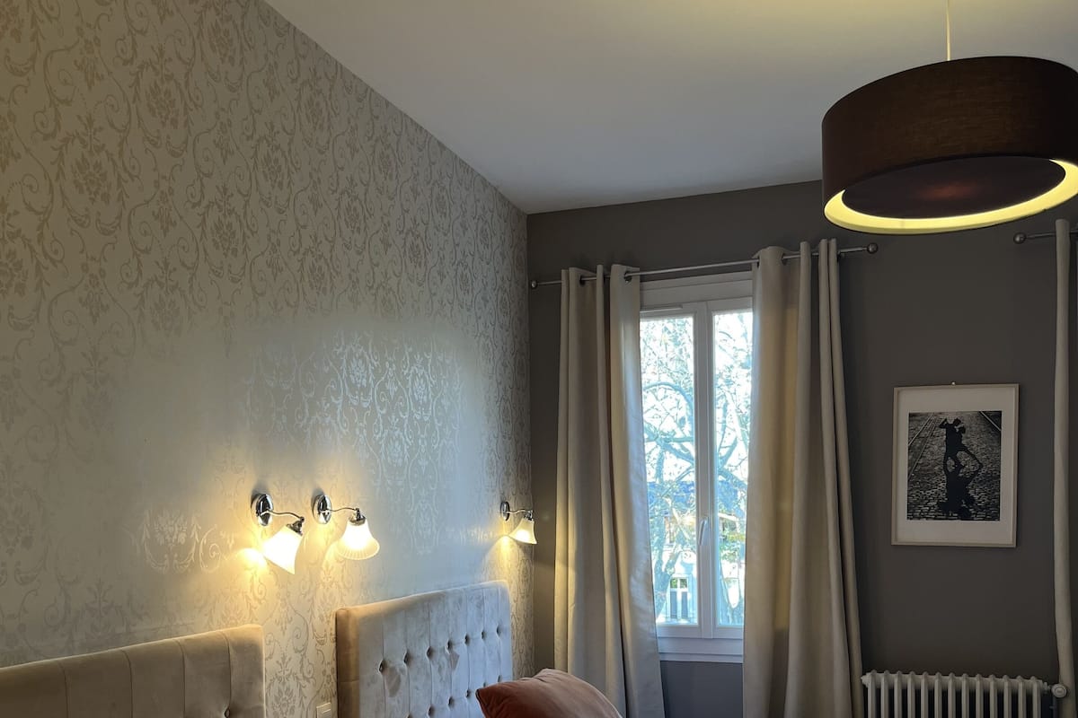 A hotel room with two beds, a large window with curtains, a radiator, and wall-mounted lights.