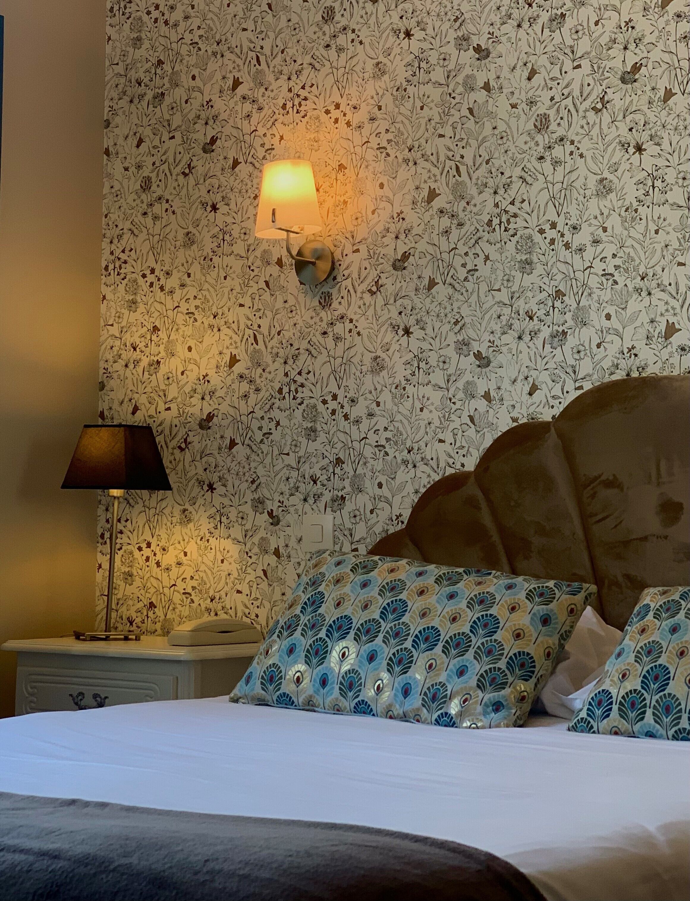 A bedroom with a floral wallpaper, a bedside lamp, a bed with patterned pillows, and a nightstand with a phone.