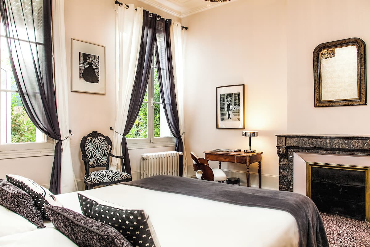 A hotel room with a bed, a chair, a desk, a mirror, and a fireplace.