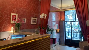 A hotel lobby with a reception desk, a chandelier, red curtains, and a large window with a view of the street. -  Hotel Mirabeau (Tours)