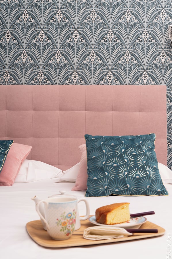Un dormitorio con un cabecero capitonado, papel tapiz con estampado floral y una cama con almohadas rosadas y turquesa. Una bandeja de madera sostiene una tetera, una taza de té, una rebanada de pastel y un libro.