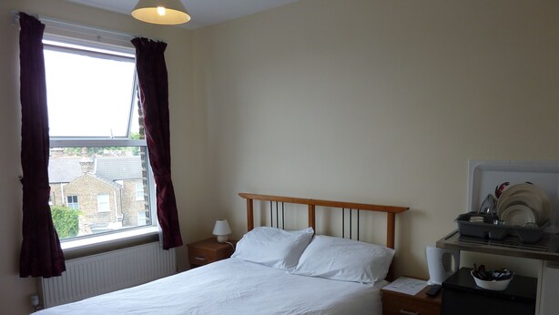 A neatly made bed with white linens, a bedside table with a lamp, and a small refrigerator. - Bennys B and B (London)
