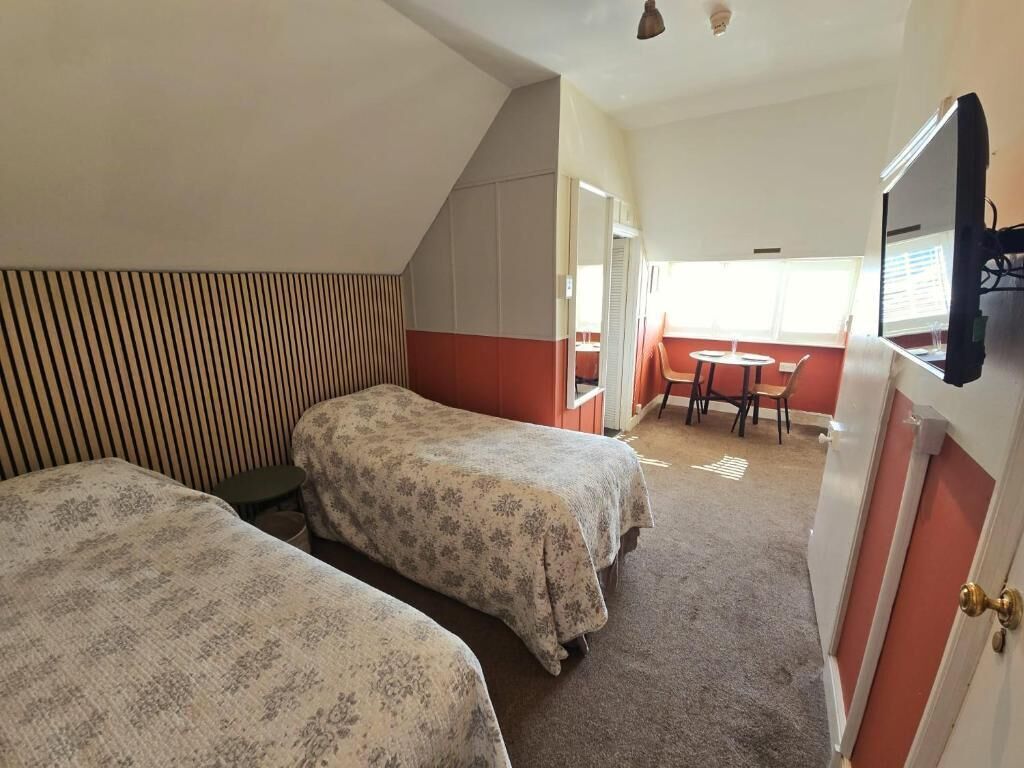 A room with two beds, a small table, and a chair. There is a door with a mirror and a light fixture above it.