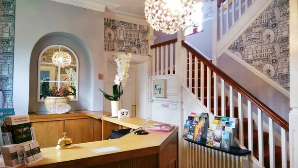 Zona de recepción de hotel con un mostrador de madera, un ventanal de arco superior y una escalera decorada con un papel pintado estampado.