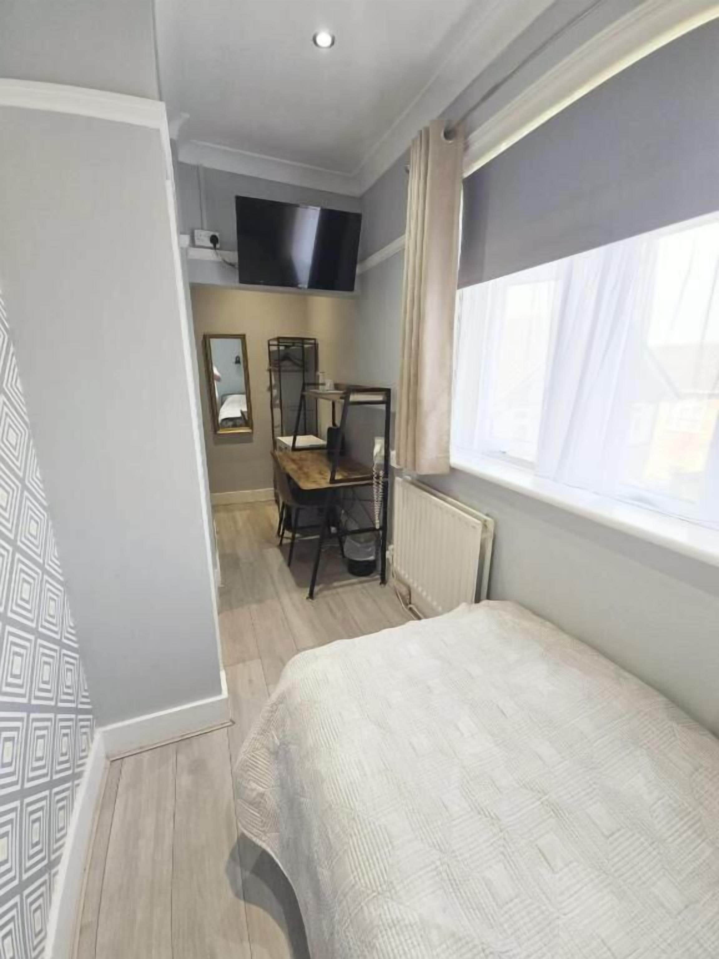Ein Schlafzimmer mit einem Bett, einem Schreibtisch, einem Sessel, einem an der Wand montierten Fernseher und einem Fenster mit Vorhängen.