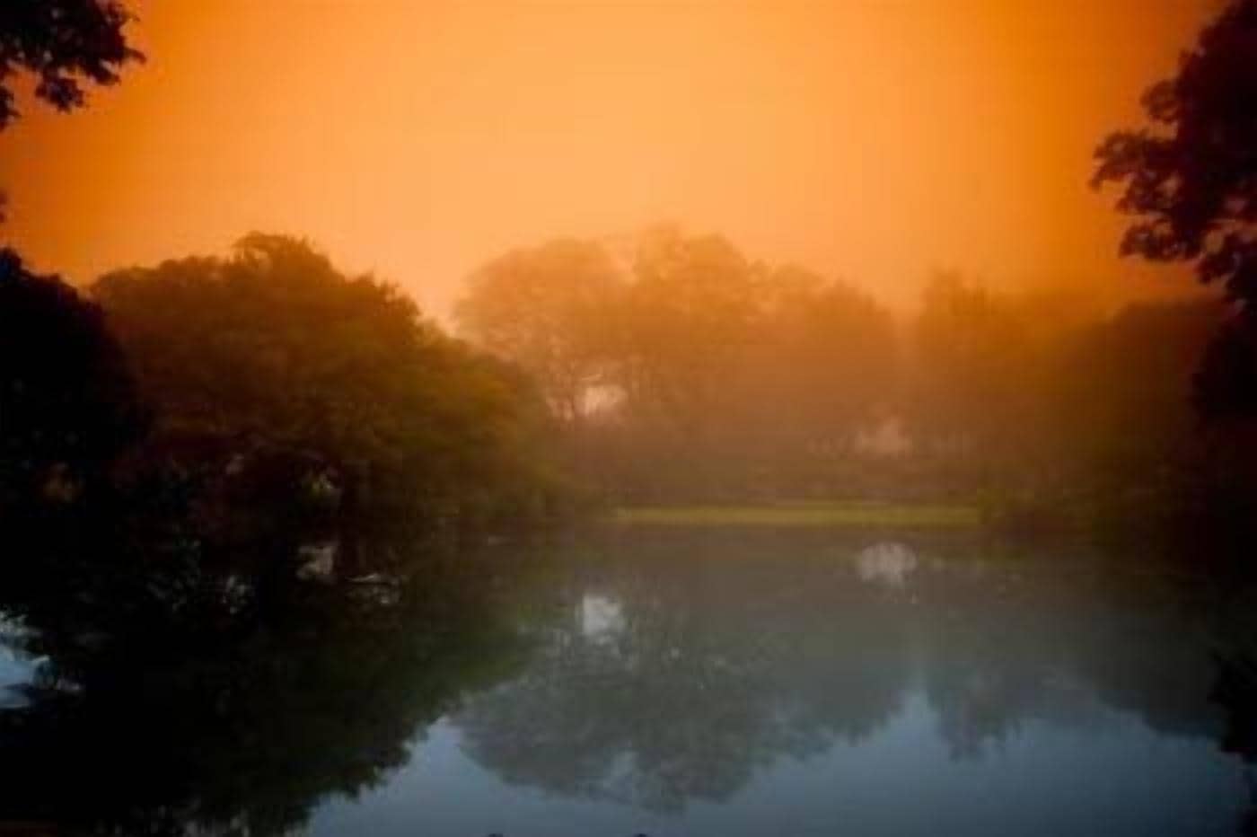 Un paysage paisible, avec une étendue d’eau qui reflète les arbres environnants et un ciel chaud aux teintes orangées.