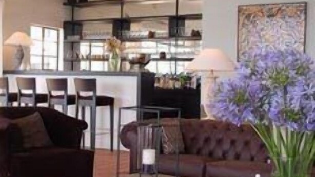 A modern living room with a dark brown sofa, a round ottoman, and a coffee table. There is a dining area with chairs and a table in the background. - Kings Walden Garden Manor (Tzaneen)